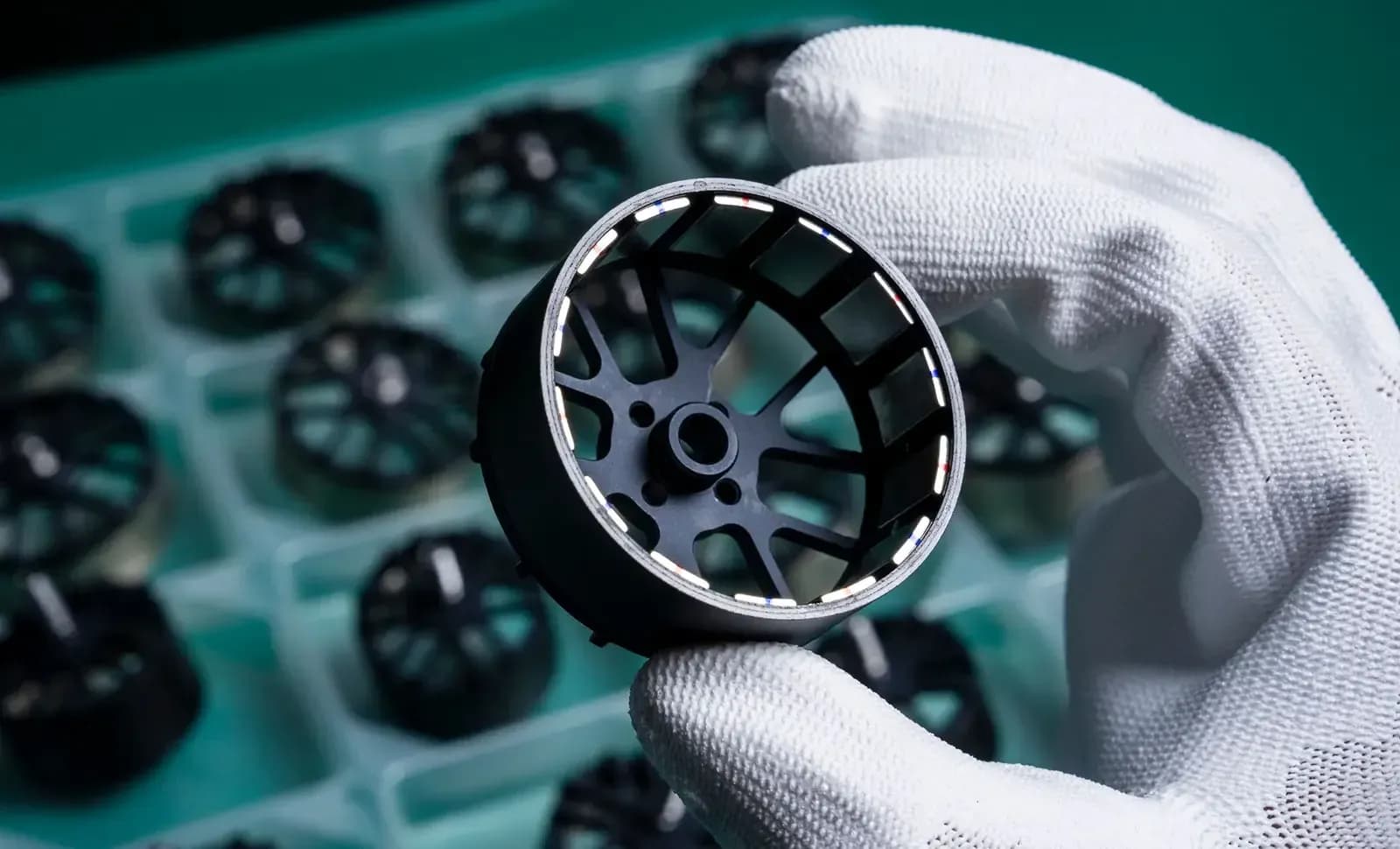 A gloved hand holds an electric motor housing above a tray of identical parts on a production line.