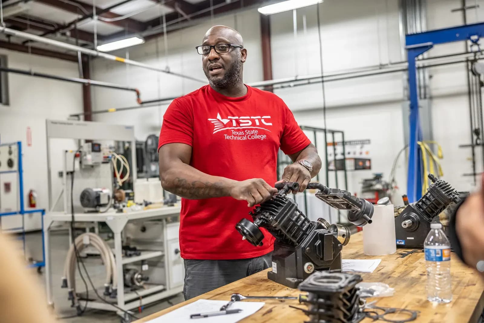 An instructor in a red Texas State Technical College shirt demonstrates a stripped compressor unit to students at a bench in the MEI training workshop in Red Oak, Texas.