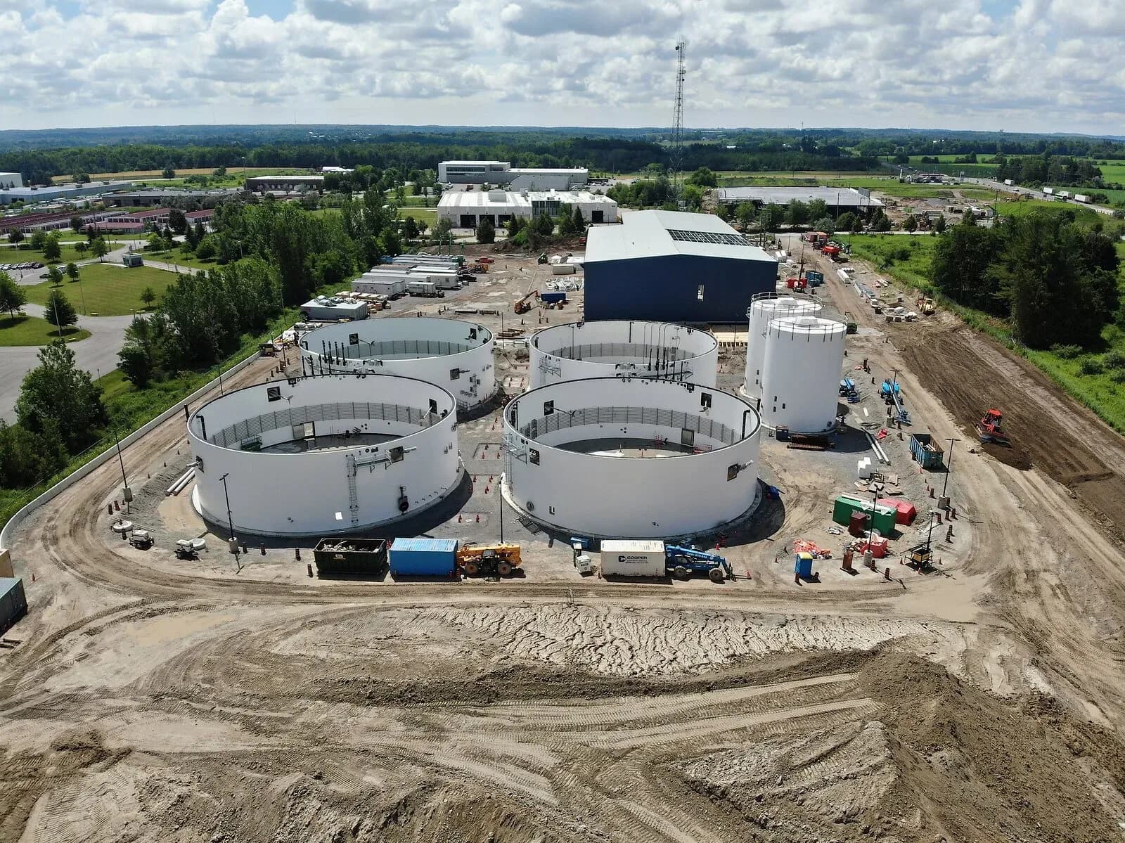Aerial view of a biogas facility under construction, with large open-topped circular digester tanks and silos beside a dark-clad industrial building on a graded site, farmland beyond.