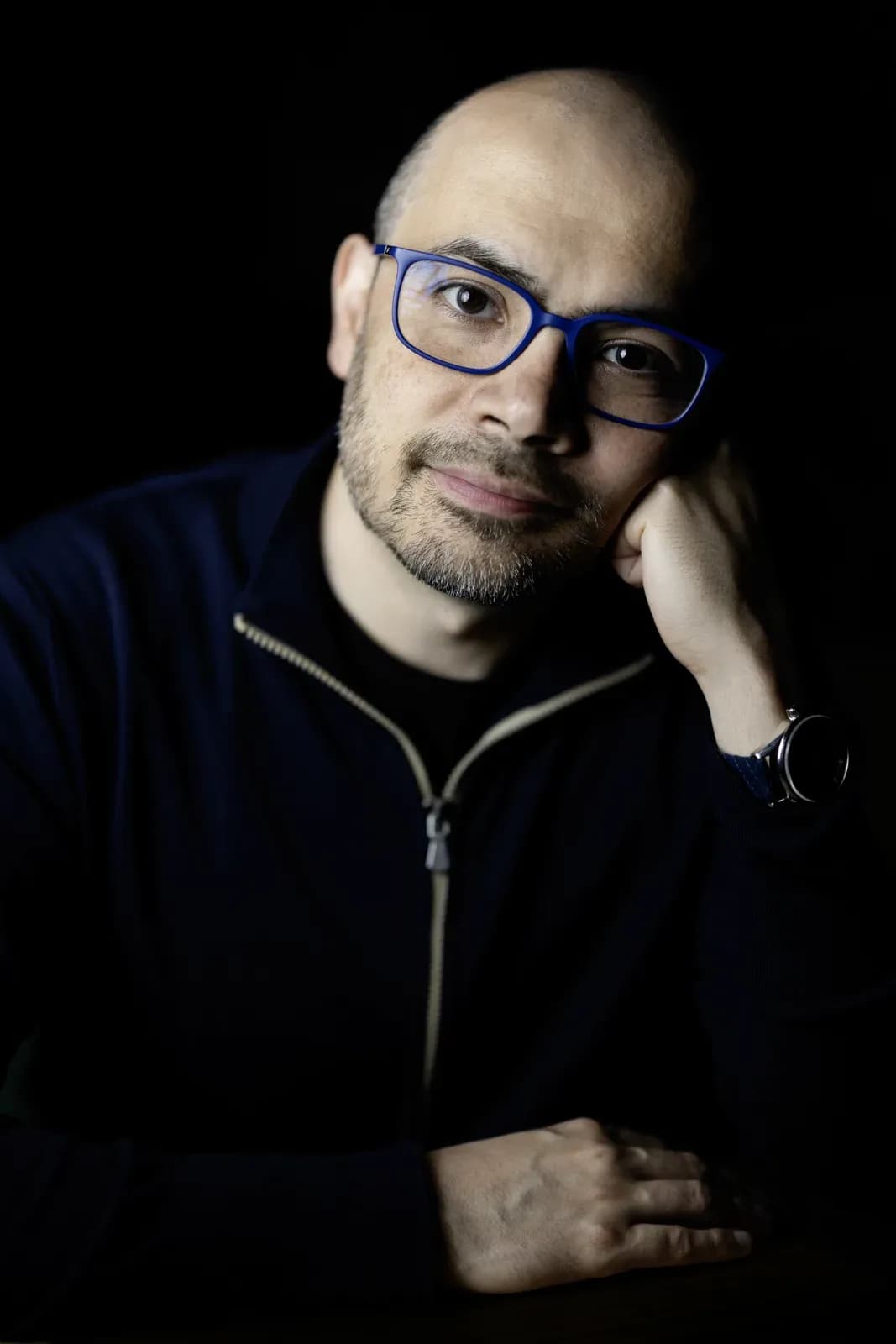 Demis Hassabis in a studio portrait, wearing glasses and a navy jacket