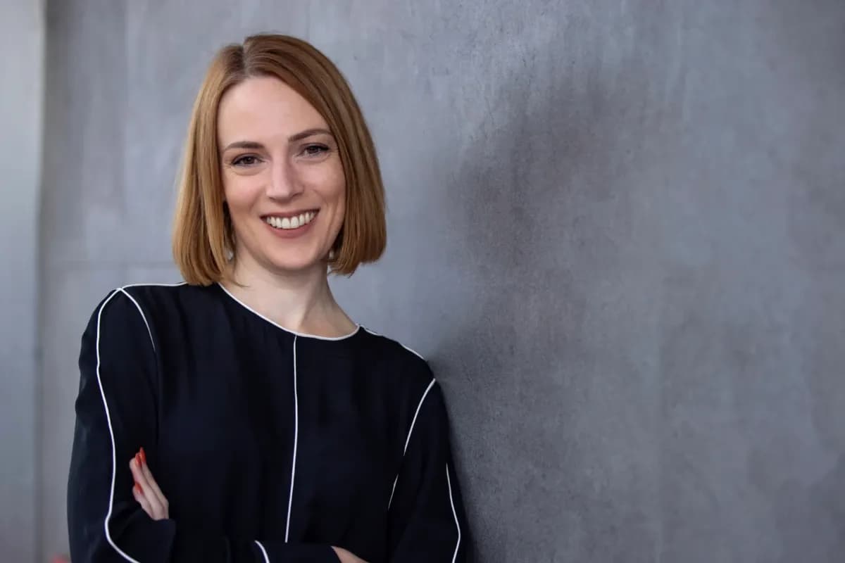 Erika Robinson standing confidently with arms crossed, wearing a modern black top with white piping detail, against a grey textured wall. She has a warm smile and shoulder-length blonde hair in a sleek bob cut.