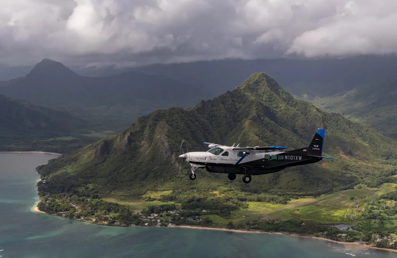 A small autonomous Joby aircraft in flight above the green mountains and coastline of Hawaii.
