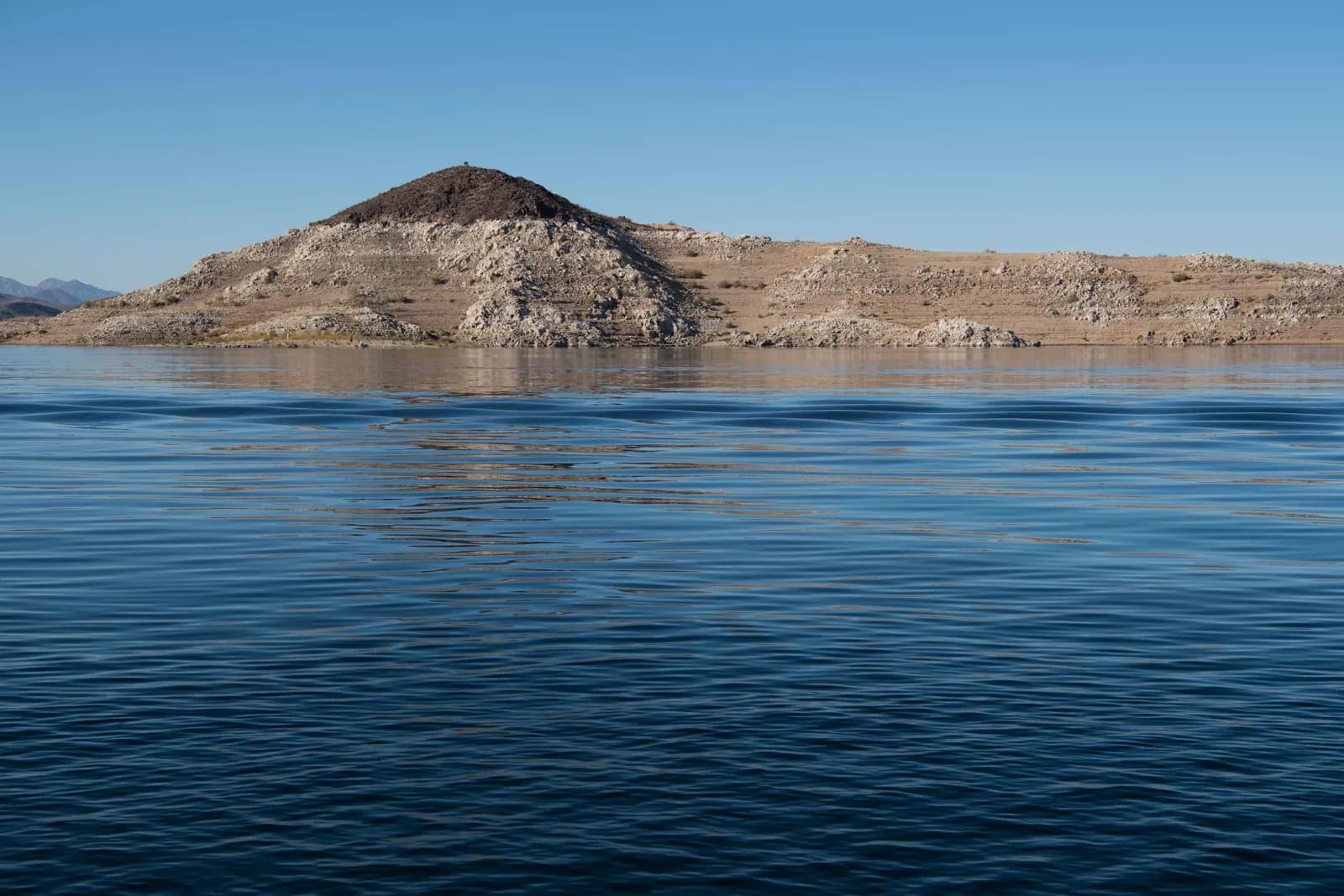 Open reservoir water at Lake Mead stretching to a dry rocky shoreline with pale mineral banding along the bank under a clear sky.