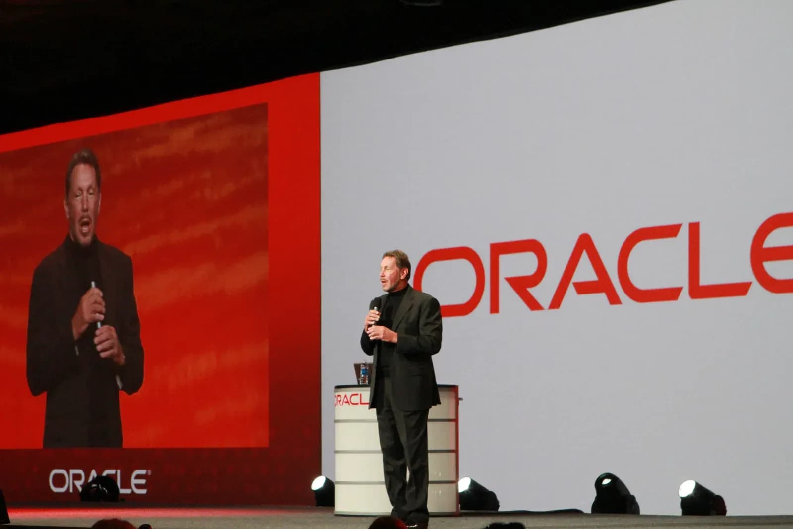 Larry Ellison speaking on stage at an Oracle OpenWorld conference, holding a microphone, with the Oracle logo on a large screen behind him