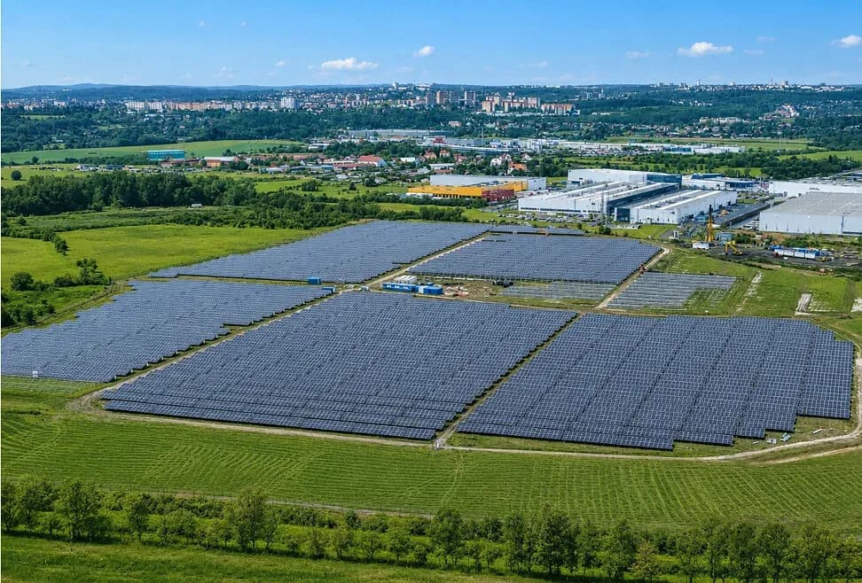 An aerial view of a large ground mounted solar farm covering reclaimed flat land on the edge of a Czech town, with industrial buildings and hills on the horizon.