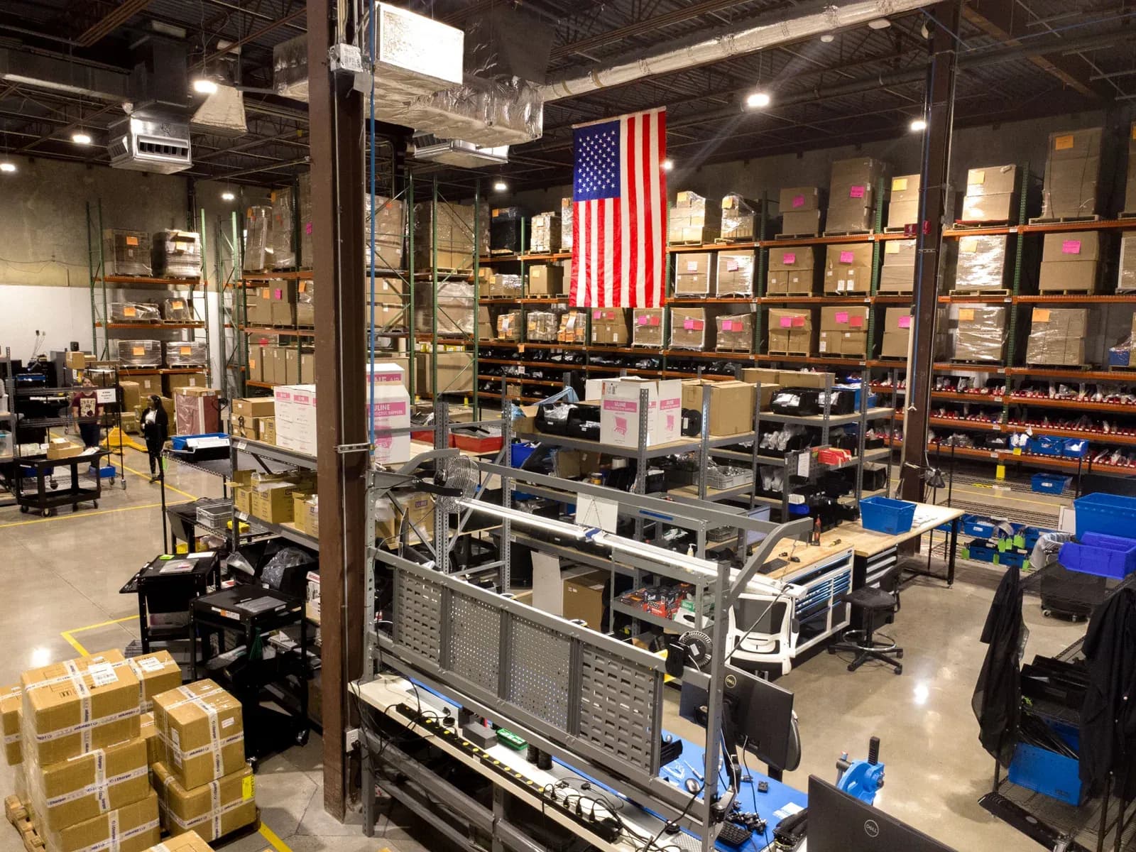 Shelves of parts and assembly benches inside the Performance Drone Works production facility in Huntsville, Alabama, with a US flag hanging above the racks