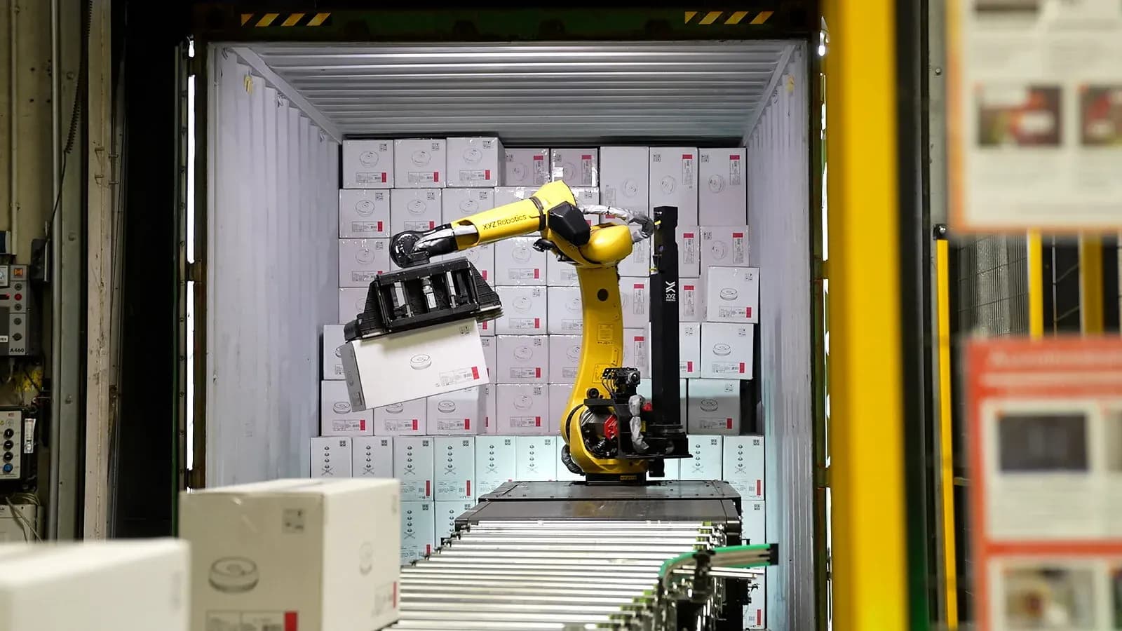 A yellow industrial robot arm reaches into a shipping container stacked with cardboard boxes and lifts one onto a roller conveyor at a warehouse loading dock.