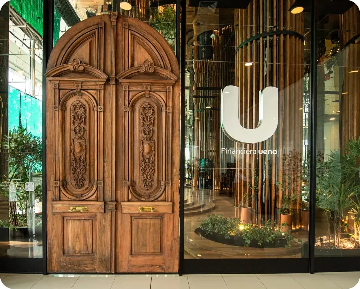 A carved wooden colonial double door standing in front of the glass entrance of a ueno branch in Paraguay, with the Financiera ueno logo on the window and a plant-filled, timber-lined lobby visible inside.