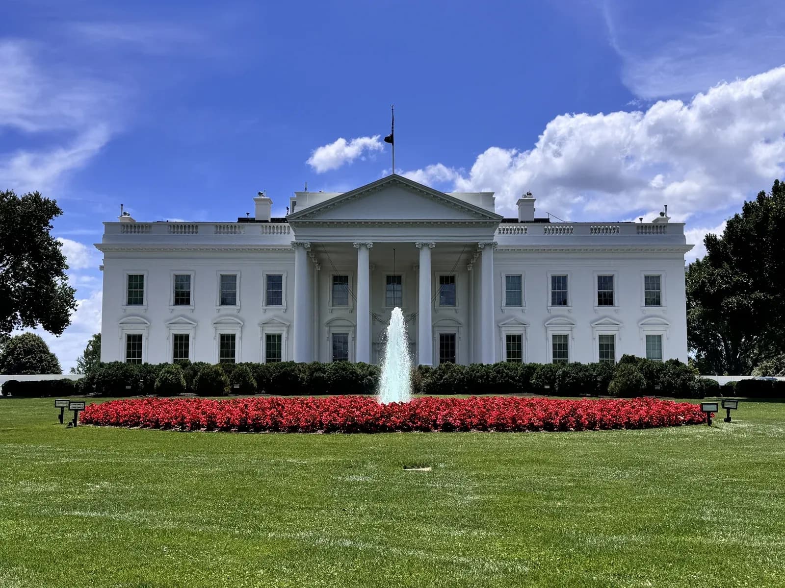 The north facade of the White House in Washington DC, seen across the North Lawn fountain and red flower beds under a blue summer sky