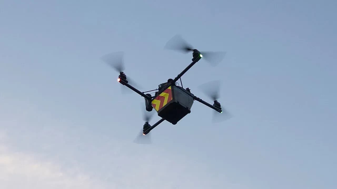 A small quadcopter drone with emergency services chevron markings in flight, carrying a boxed medical payload slung beneath it.