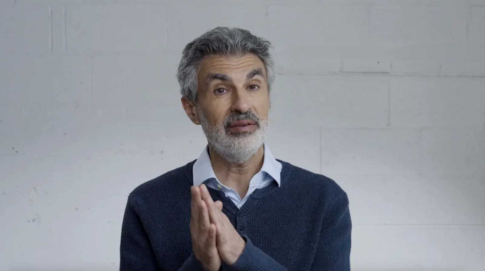 Yoshua Bengio speaking to camera, hands clasped, against a white brick wall