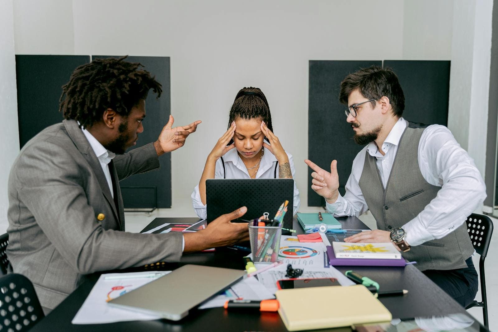 Two men argue while a woman looks frustrated at a laptop in an office environment.