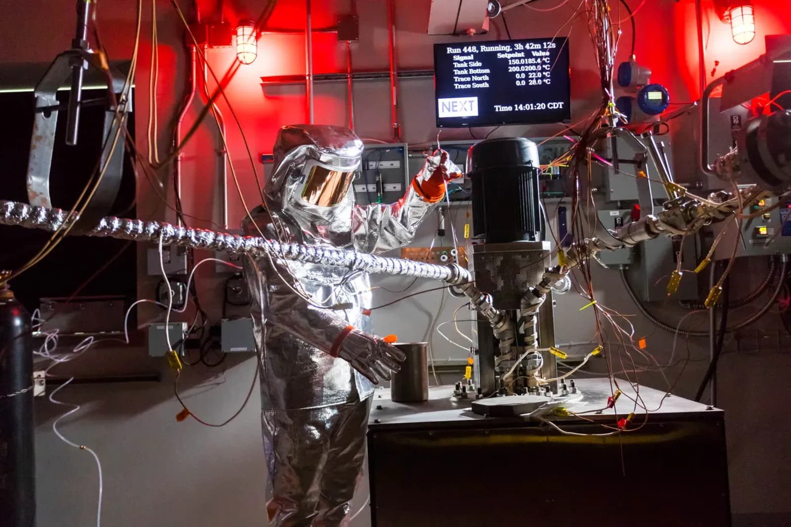 A researcher in a silver heat-protective suit works at a molten-salt test loop in Abilene Christian University's NEXT Lab, with a run-status monitor overhead.
