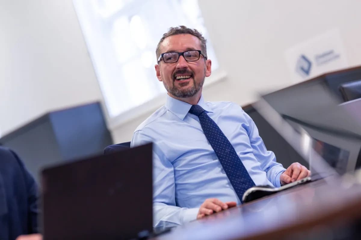 A professional man in a light blue shirt and navy polka dot tie, wearing glasses, smiles warmly while seated at his desk. He is photographed from a slightly lower angle, giving a confident and approachable impression.