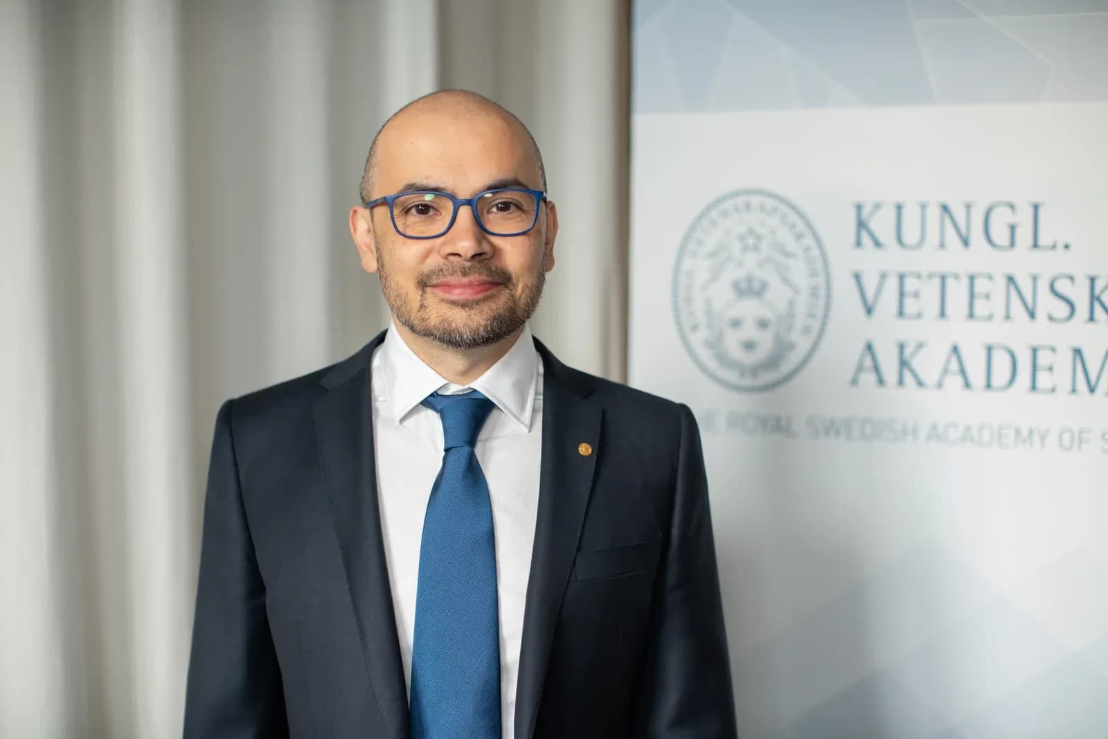 Demis Hassabis on stage at the 2024 Nobel Prize events, seated in an armchair speaking, wearing a dark suit