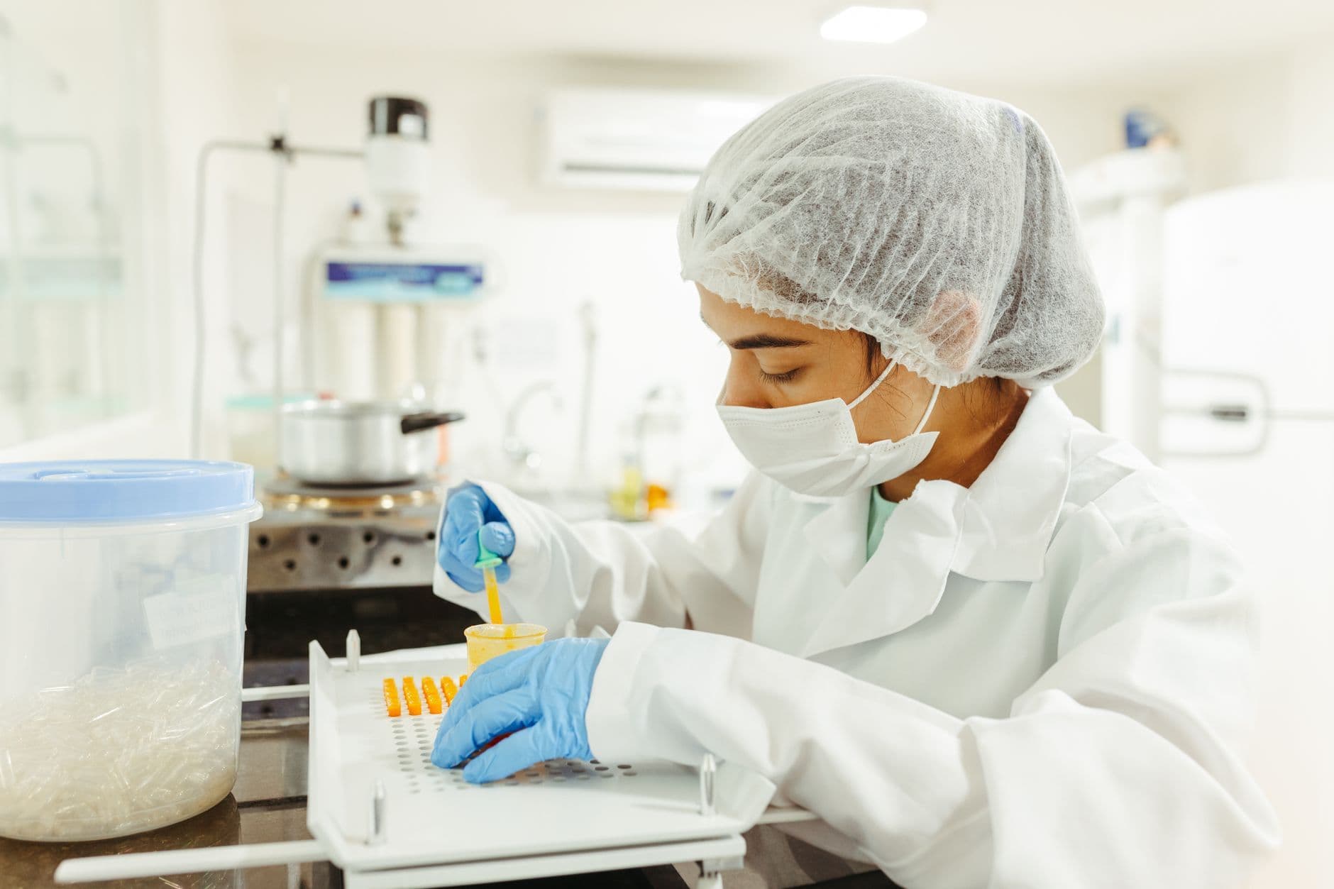 A pharmaceutical worker in a hairnet, mask and gloves filling capsules at a manufacturing bench