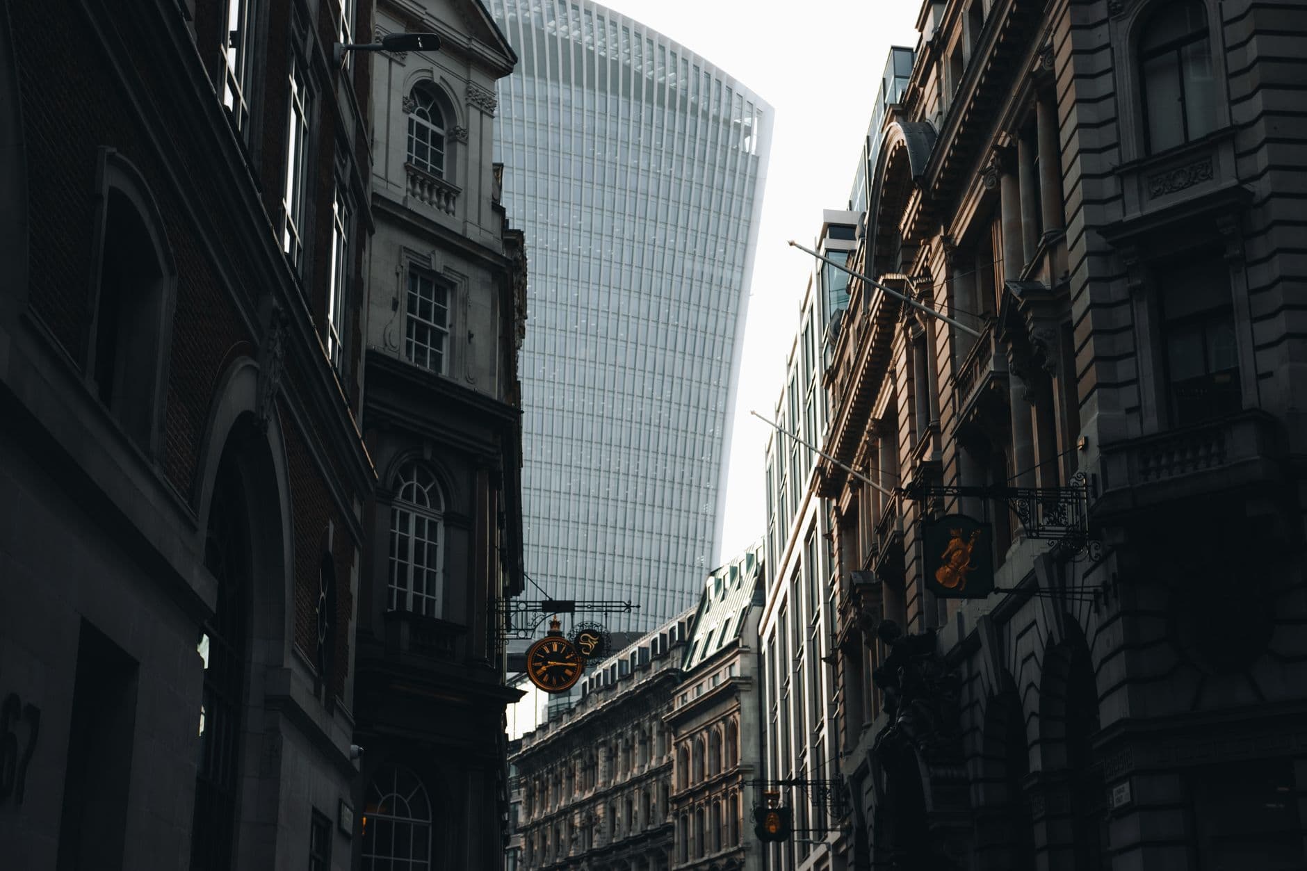 A modern glass tower rises between older stone buildings on a street in the City of London
