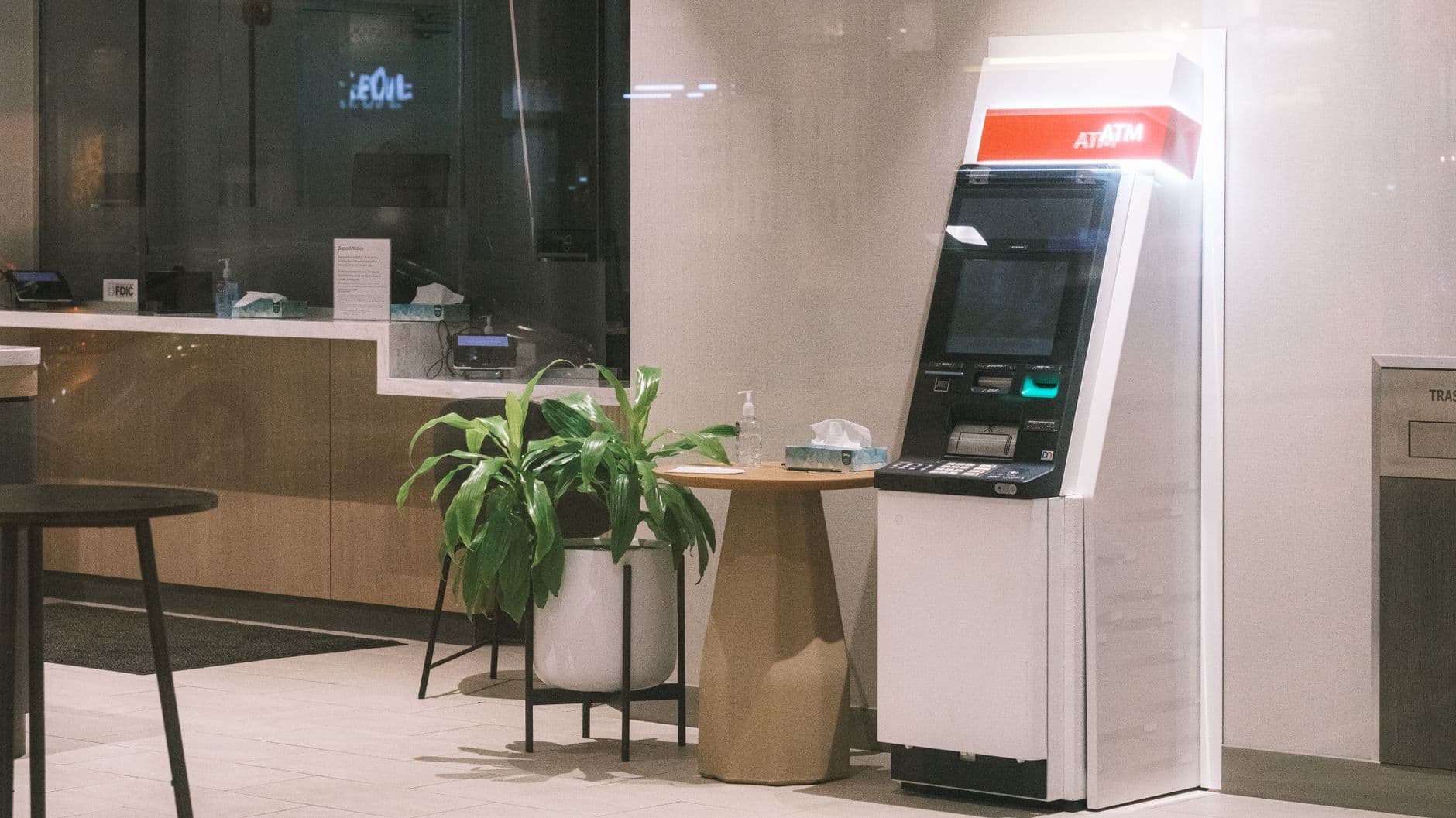 An empty bank lobby with a teller counter behind glass and a freestanding ATM