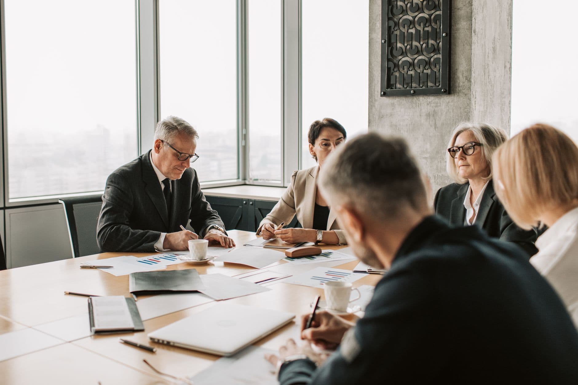 Senior executives in a boardroom reviewing printed financial documents with a city skyline behind them