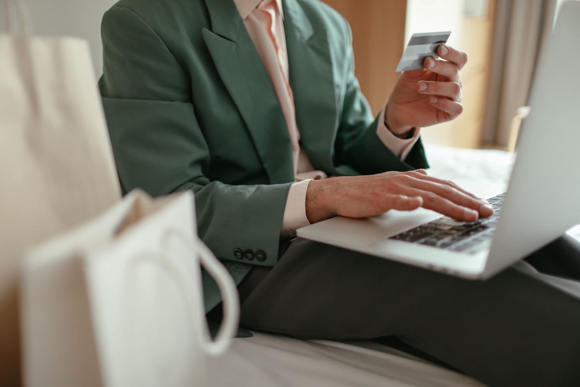 A person in a blazer holds a credit card while shopping on a laptop, with shopping bags nearby