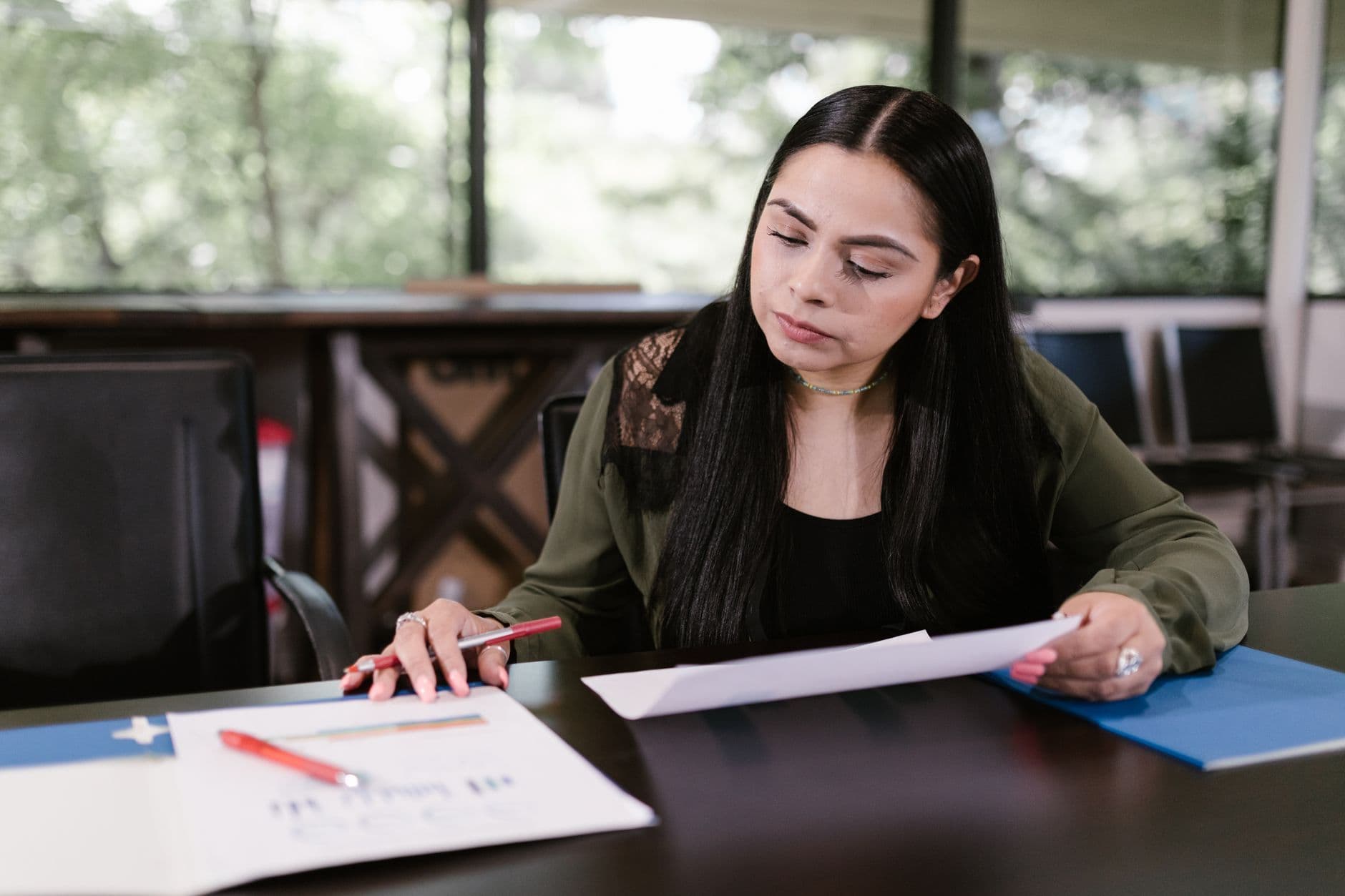 A woman at an office desk reads a printed document with a pen in hand, charts on the table in front of her