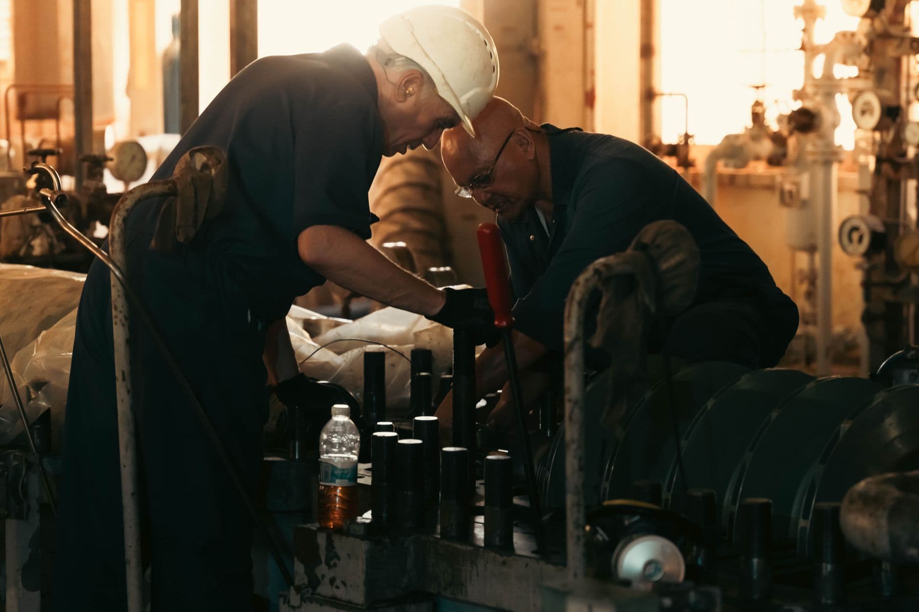 Two workers in overalls lean over machinery together on an industrial plant floor