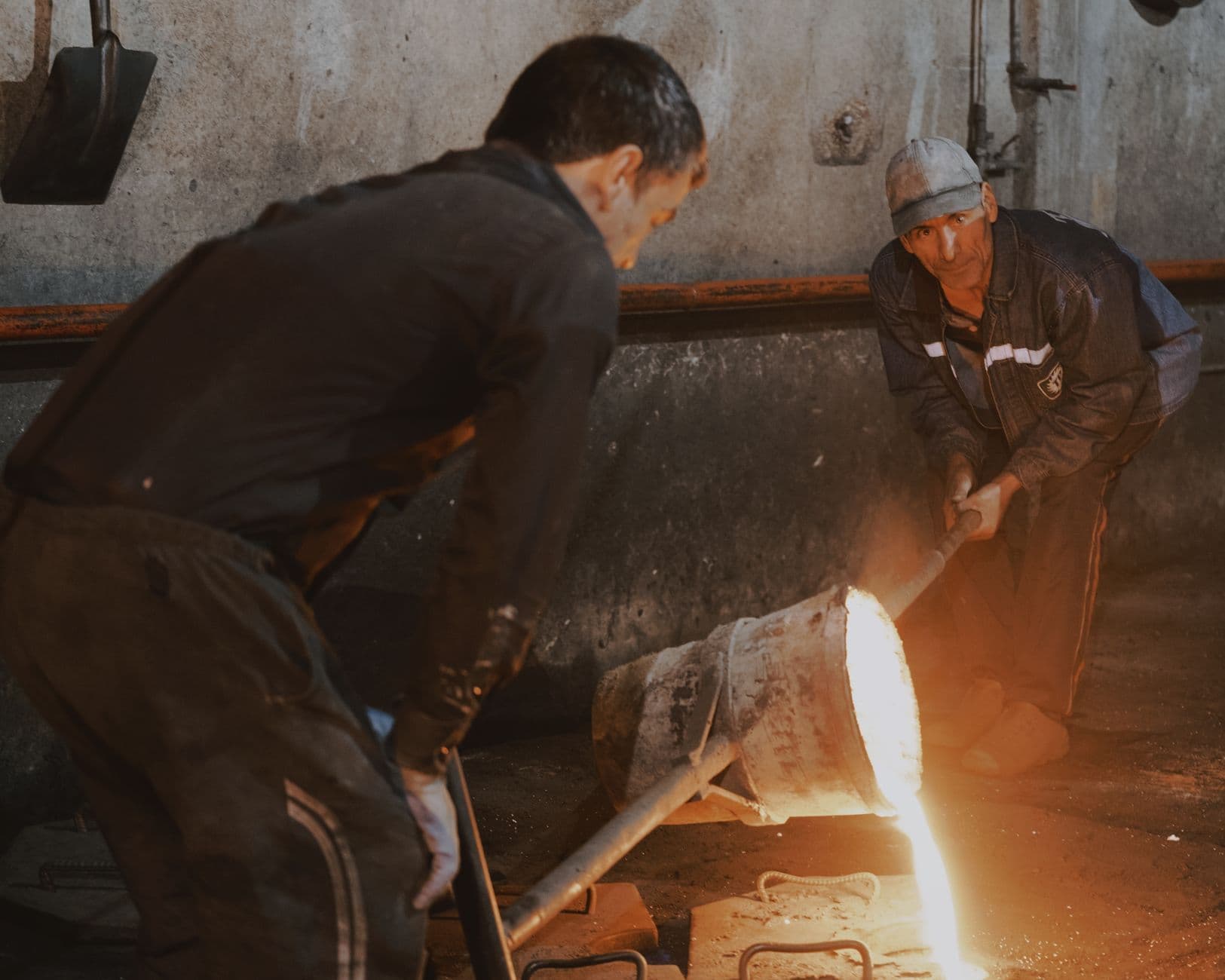 Two foundry workers pour molten metal from a ladle into a casting mould