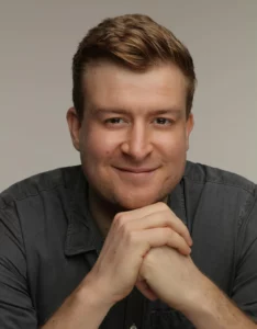 A headshot of a young man with brown hair styled upward, wearing a dark grey collared shirt, smiling warmly at the camera with his hands clasped under his chin. The image has a neutral grey background and professional studio lighting.