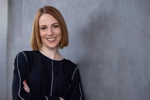 Erika Robinson standing confidently with arms crossed, wearing a modern black top with white piping detail, against a grey textured wall. She has a warm smile and shoulder-length blonde hair in a sleek bob cut.