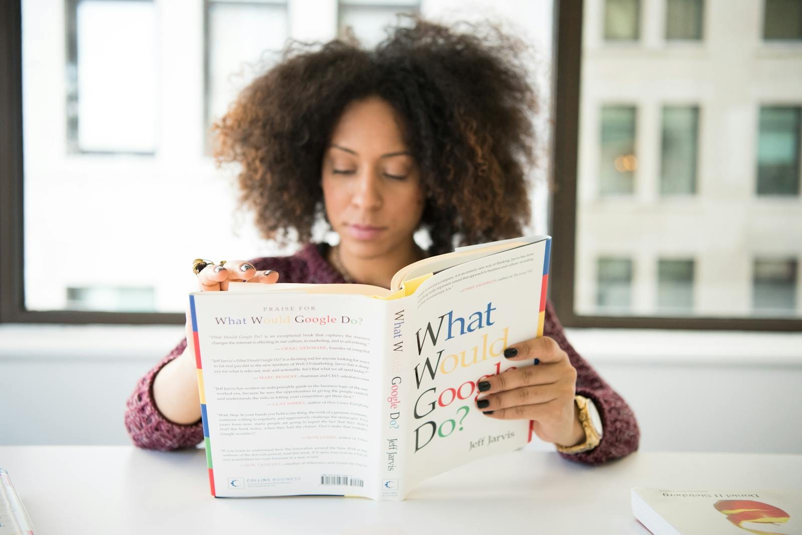 Google Finally Gets It Right with Workspace Gemini Integration - Well, Almost 1 A woman reading 'What Would Google Do?' at a desk by a window in a modern office.