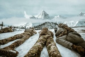 Soldiers in camouflage lying in snow during winter training in mountain landscape.