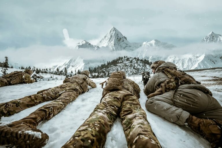 Soldiers in camouflage lying in snow during winter training in mountain landscape.