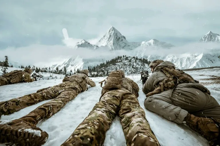 Soldiers in camouflage lying in snow during winter training in mountain landscape.